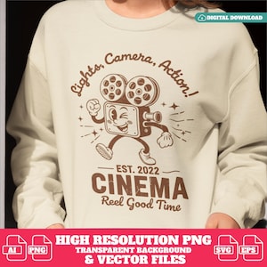 Könnte beinhalten: Beiges Sweatshirt mit braunem Vintage-Kino-Motiv. Das Design zeigt eine Cartoon-Kamera mit dem Text "Lights, Camera, Action!" und "Cinema Reel Good Time".