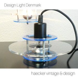 May include: A modern, minimalist table lamp with a clear acrylic base and a black cord. The lamp has two circular, clear acrylic shades with blue trim. The lamp is lit with a light bulb.