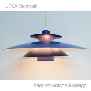 May include: A blue and white pendant light with a layered, saucer-shaped design. The light is hanging from a white cord and is turned on, illuminating the room. The text "JEKA Denmark" is visible in the top left corner of the image.