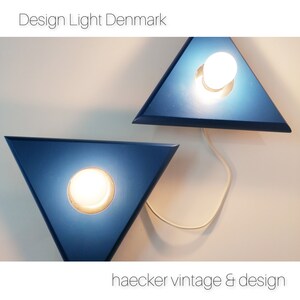 May include: Two blue triangular wall sconces with white light bulbs. The sconces are connected by a white cord. The text "Design Light Denmark" is at the top of the image and "haecker vintage & design" is at the bottom.