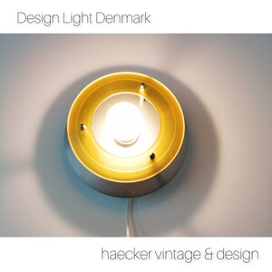 May include: A round, vintage wall light fixture with a silver base and a gold-colored inner ring. The light has a bright bulb in the center and is mounted on a white wall. The text "Design Light Denmark" and "haecker vintage & design" are visible.