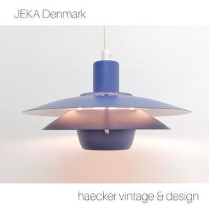 May include: A blue and white pendant light with a circular, saucer-shaped shade. The light is suspended from a white cord and has a perforated metal design on the top of the shade. The text "JEKA Denmark" is visible at the top of the image.
