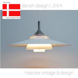 May include: A white, saucer-shaped pendant light with a silver metal frame and a small, round, white light fixture. The light is hanging from a silver cord. The text "danish design | JEKA" is visible at the top of the image.