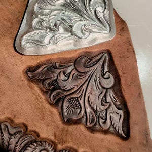 Op de afbeelding: Een stuk gelooid leer met drie verschillende bloemmotieven in het oppervlak gedrukt. Eén motief is een glanzende zilveren metalen plaat, terwijl de andere twee diep in het leer zijn gestempeld met donkere, schaduwrijke details.
