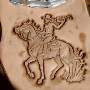 Könnte beinhalten: Ein hellbraunes Lederstück mit einem detaillierten geprägten Bild eines Cowboys auf einem Pferd, der einen Lasso hält. Ein Metallstempel eines Cowboys auf einem Pferd ist oben sichtbar. Der Cowboy trägt einen Hut und wirft gerade das Lasso.