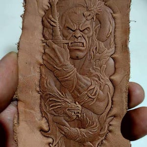 May include: A detailed, hand-tooled leather piece depicting a fierce warrior with a sword, flanked by two dragons. The artwork is in shades of brown, showcasing intricate relief carving and a textured surface. The warrior's face is contorted in a grimace.