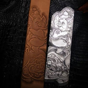 Peut inclure: Une ceinture en cuir avec un motif en relief détaillé d'un cochon portant un chapeau et debout sur des nuages. Le motif est présenté à la fois en cuir brun et en métal argenté.
