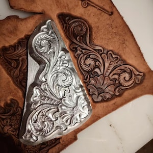 May include: A silver-colored metal stamping tool with an intricate floral and scroll pattern, positioned over a piece of tan leather that features a matching embossed floral design and a small stamped airplane icon at the top edge.