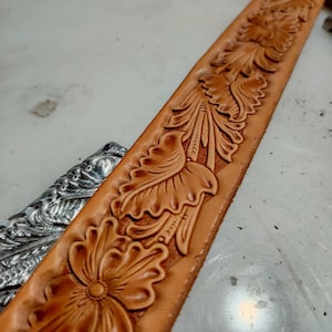 Può includere: Una cinghia per cintura in pelle color cuoio con un motivo floreale in rilievo continuo con petali e foglie dettagliati. Uno strumento in metallo color argento con un intricato design inciso è posizionato parzialmente sotto la cinghia in pelle.