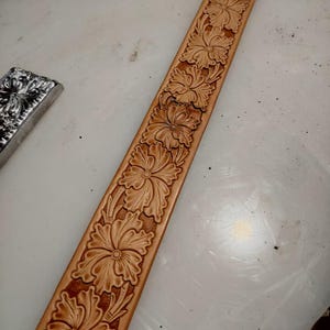 Könnte beinhalten: Ein beigefarbener Ledergürtelriemen mit einem durchgehenden geprägten Blumenmuster mit detaillierten Blütenblättern und Blättern. Das Leder hat eine glatte, natürliche Oberfläche und das florale Design ist tief eingeprägt, was eine Textur erzeugt.