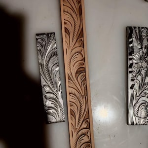 May include: A long, light tan leather strip featuring a deeply embossed, intricate floral and leaf pattern with dark brown shading. Beside it are two rectangular metal stamping tools, each displaying a matching relief design of leaves and petals.