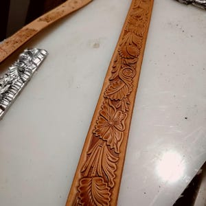 May include: A long, tan leather belt strap featuring a continuous, deeply embossed floral and leaf pattern along its entire length. The tooling displays intricate petals, swirling foliage, and textured backgrounds, showcasing natural tan leather tones.