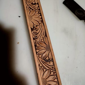 May include: A tan leather belt strap featuring a continuous embossed pattern of sunflowers and swirling leaves. The floral design is deeply tooled into the surface, creating a textured appearance with dark brown accents within the recessed areas.