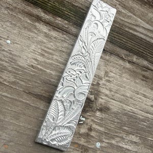 Pu&ograve; includere: Un oggetto metallico argentato di forma rettangolare con intricati motivi floreali e di foglie scolpiti sulla sua superficie. Il design dettagliato presenta linee a spirale ed elementi testurizzati, creando un aspetto decorativo e ornato. L'oggetto &egrave; posizionato diagonalmente su una superficie di legno invecchiato.