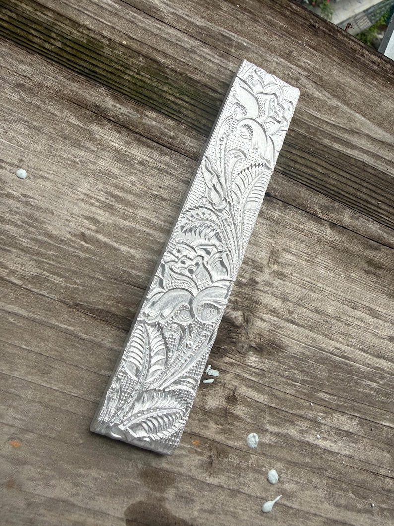 Pu&ograve; includere: Un oggetto metallico argentato di forma rettangolare con intricati motivi floreali e di foglie scolpiti sulla sua superficie. Il design dettagliato presenta linee a spirale ed elementi testurizzati, creando un aspetto decorativo e ornato. L'oggetto &egrave; posizionato diagonalmente su una superficie di legno invecchiato.