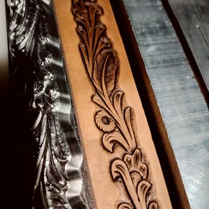 May include: A tan leather strip featuring a deeply embossed, continuous floral scroll pattern with dark brown shading, positioned between two metallic, silver-coloured steel stamping tools with matching intricate carved designs.