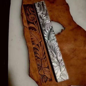 May include: A rectangular metal stamping tool with a detailed floral and leaf pattern embossed on its surface, positioned next to a strip of brown leather that features the same intricate floral design pressed into the material.
