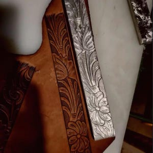 May include: A tan leather strip featuring a deeply embossed floral and leaf pattern with dark brown highlights, positioned next to a metallic silver strip that displays an identical intricate botanical design in relief.