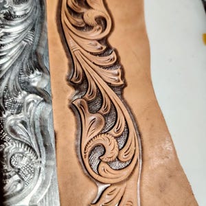 May include: A piece of tan leather featuring a deeply embossed and carved floral scroll pattern. The design includes intricate swirling leaves and petals with a textured, cross-hatched background, showcasing the depth of the tooling work.