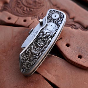 Könnte beinhalten: Ein Klappmesser mit einem silberfarbenen Metallgriff, der eine detaillierte Gravur eines Schädels umgeben von kunstvollen Schnörkeln aufweist. Die Klinge ist teilweise sichtbar und verfügt über einen kleinen Daumenpin sowie ein Pfeilsymbol.