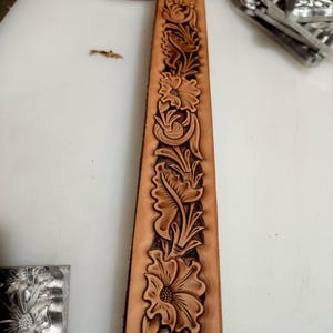 May include: A long, tan leather belt strap featuring a continuous, deeply embossed floral pattern. The design consists of intricate, repeating flower blossoms and leafy vines carved into the surface, creating a textured, three-dimensional effect.