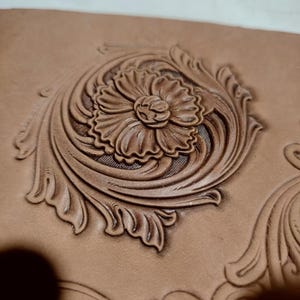 Könnte beinhalten: Nahaufnahme eines detaillierten, handgearbeiteten Lederdesigns. Der Fokus liegt auf einer zentralen Blume mit geschichteten Blütenblättern, umgeben von wirbelnden Blättern und Ranken. Die Prägung erzeugt einen 3D-Effekt und zeigt die Handwerkskunst. Das Leder hat eine warme, natürliche, hellbraune Farbe.