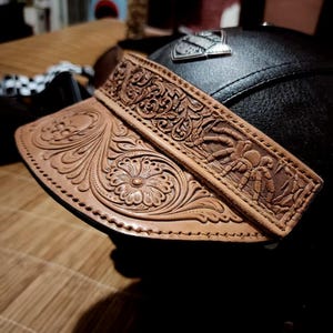 May include: A black leather motorcycle helmet featuring a tan leather visor attachment. The visor is decorated with intricate hand-tooled floral patterns and a detailed spider motif. A small silver-toned metal emblem is attached to the helmet crown.