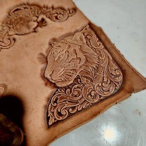 May include: A piece of light tan leather featuring a detailed embossed tiger head profile surrounded by intricate swirling floral scrollwork patterns. The design is deeply stamped into the surface, creating a textured relief effect on the material.