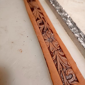 Puede incluir: Una tira de cuero color canela con un patrón floral y de volutas profundamente grabado junto a una herramienta de estampado de metal plateado a juego. El cuero muestra intrincados detalles tallados de pétalos y hojas, mientras que la herramienta muestra el diseño inverso.