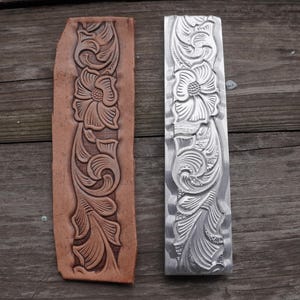 Könnte beinhalten: Ein rechteckiges Stück lohfarbenes Leder mit einem tief geprägten Blumen- und Rankenmuster neben einem passenden silberfarbenen Metallstempelwerkzeug, das dasselbe komplexe botanische Design auf seiner Oberfläche eingraviert hat.
