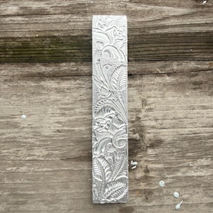 Pu&ograve; includere: Un oggetto rettangolare in metallo color argento con un intricato motivo floreale. Il design presenta foglie dettagliate, viti e altri elementi botanici. L'oggetto &egrave; esposto su una superficie di legno stagionato.