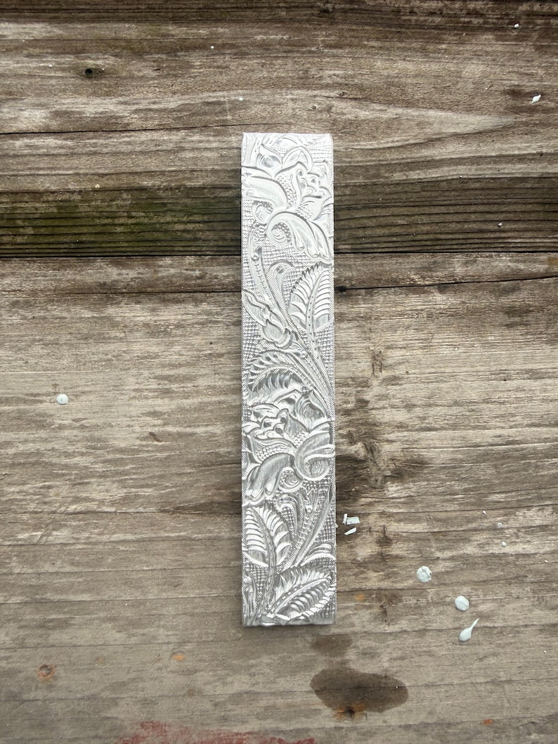 Pu&ograve; includere: Un oggetto rettangolare in metallo color argento con un intricato motivo floreale. Il design presenta foglie dettagliate, viti e altri elementi botanici. L'oggetto &egrave; esposto su una superficie di legno stagionato.