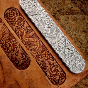 Puede incluir: Primer plano de una pieza de cuero con dos correas de cuero tallado y una correa de metal plateado. Las correas de cuero son marrones con intrincados diseños florales y geométricos. La correa plateada tiene grabados detallados.