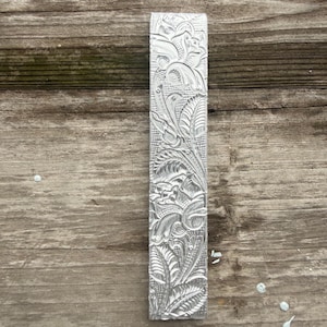 Pu&ograve; includere: Un oggetto rettangolare in metallo color argento con un intricato motivo floreale. Il design presenta foglie, fiori ed elementi a spirale dettagliati. L'oggetto &egrave; esposto su una superficie di legno invecchiato.
