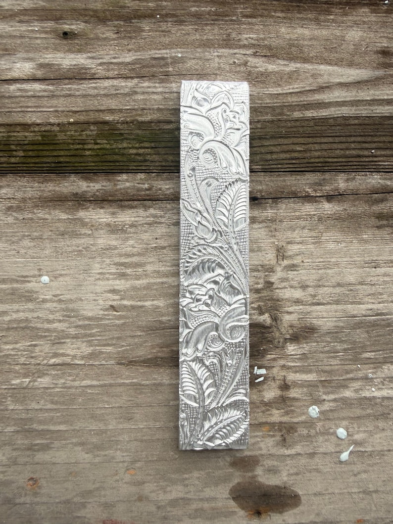 Pu&ograve; includere: Un oggetto rettangolare in metallo color argento con un intricato motivo floreale. Il design presenta foglie, fiori ed elementi a spirale dettagliati. L'oggetto &egrave; esposto su una superficie di legno invecchiato.