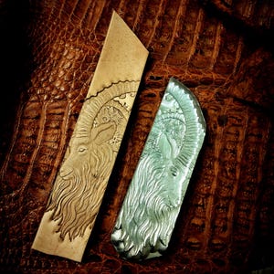 May include: Two rectangular objects with engraved ram designs. One is tan, the other is silver. The ram's head is detailed with curved horns and a long beard. The background is a textured brown surface.