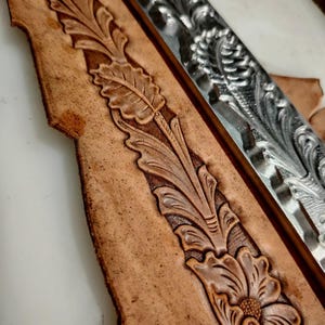 May include: A tan leather strip featuring a deeply embossed floral and leaf pattern running vertically, positioned next to a long, silver-colored metal stamping tool that displays the same intricate botanical design in a raised relief format.