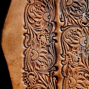 Puede incluir: Primer plano de cuero tallado intrincadamente, que muestra un patrón floral. El diseño presenta un trabajo de relieve detallado de flores y hojas estilizadas, creando un efecto texturizado y tridimensional. El cuero tiene un tono marrón cálido y natural.