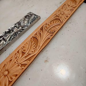 Peut inclure: Une longue bande de cuir beige présentant un motif floral et végétal profondément gaufré, accompagnée d'un outil d'estampage en métal argenté assorti. Le cuir affiche des détails botaniques sculptés avec des zones en creux assombries.
