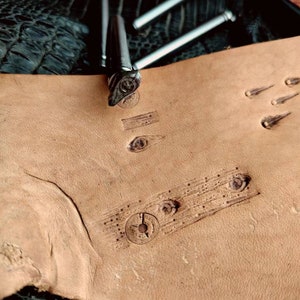 Op de afbeelding: Een close-up van een leerambachtelijk project met verschillende leerstempels. De stempels worden gebruikt om ontwerpen op leer te maken. Het leer is lichtbruin van kleur en de stempels zijn van metaal.