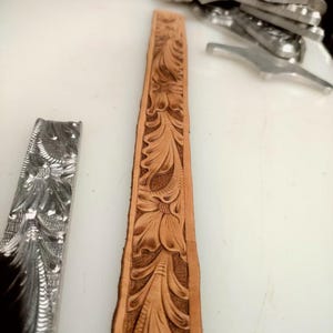 May include: A long, tan leather strip featuring an intricate, hand-tooled floral and leaf pattern. Beside it lies a silver-colored metal stamping tool with an identical embossed design, used for creating the detailed impressions on the leather surface.