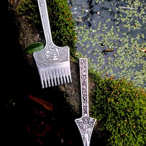 May include: Two ornate silver serving utensils. One is a comb-like tool with a detailed handle and a decorative face. The other is a three-pronged fork with a detailed handle and a skull design. Both are set against a backdrop of moss and water.