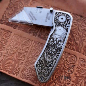 May include: A folding utility knife with a stainless steel blade and a silver-toned handle featuring an intricate engraved skull design with ornate scrollwork. The blade is partially extended and displays the text STAINLESS STEEL.