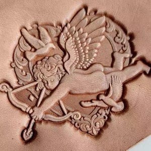 May include: A detailed relief carving of Cupid, complete with bow and arrow, wings, and a dove, set within an ornate frame. The artwork is embossed on a light brown leather surface, showcasing intricate details and textures. The design is likely for leathercraft.