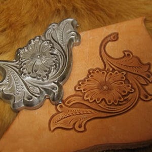 Puede incluir: Un sello de metal plateado con diseño floral junto a un trozo de cuero marrón claro con un patrón floral en relieve a juego. El sello es muy detallado y el cuero muestra la impresión del diseño.