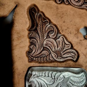 May include: A tan leather piece featuring a deeply embossed floral and scroll pattern, with a silver metal stamping tool positioned at the bottom, showing the matching intricate design used to create the impression in the leather surface.