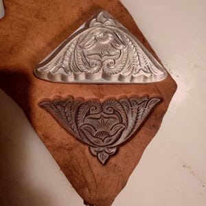 Peut inclure: Un outil de marquage en métal triangulaire présentant un motif floral et de volutes complexe, placé au-dessus d'un morceau de cuir beige qui affiche une empreinte profonde et nette du même motif orné estampé sur sa surface.