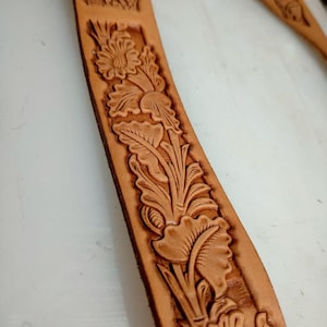 May include: A tan leather belt strap featuring a continuous embossed floral pattern. The design consists of detailed flowers and leaves pressed into the surface of the light brown leather, creating a textured, repeating botanical motif along the length.