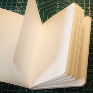 Könnte beinhalten: Ein weißes, leeres Notizbuch mit strukturiertem Einband ist auf einer leeren Seite geöffnet. Das Notizbuch liegt auf einer grünen Schneidematte.