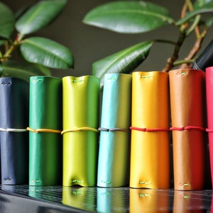 Könnte beinhalten: Eine Sammlung farbenfroher Leder-Notizbuchhüllen in verschiedenen Farbtönen, darunter Grau, Blau, Grün, Gelb, Orange und Rot. Jede Hülle ist aufgerollt und mit einem Gummiband befestigt. Die Notizbücher sind in einer Reihe angeordnet und zeigen ein Farbspektrum.